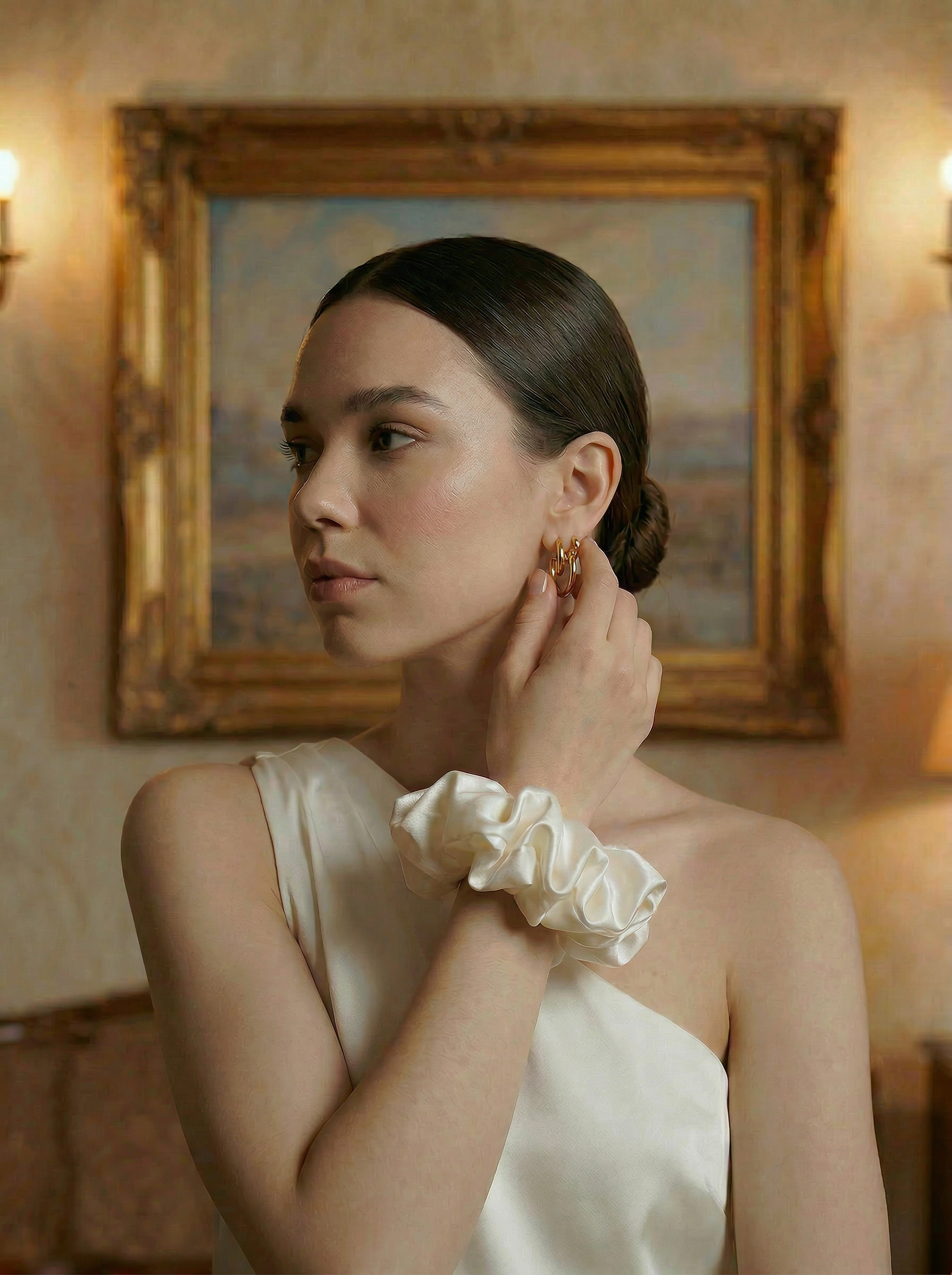 Woman adjusting her earring in a room with a painting on the wall she's wearing an ivory silk crunchie on her wrist